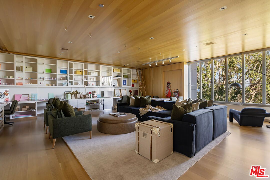 This is a spacious living room featuring a wall-to-wall built-in shelving unit, a large sectional sofa with olive green pillows, and a round ottoman. The room is well-lit with natural light from a large window and has a warm, inviting atmosphere with hardwood floors and a light-colored area rug. A decorative trunk sits in the foreground, adding a touch of elegance.