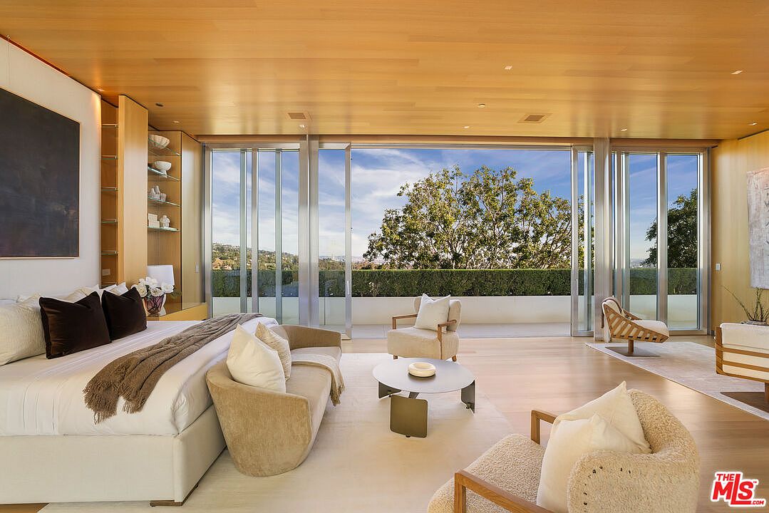 This is a luxurious primary bedroom featuring a large bed with plush pillows and a throw blanket. The room has a light wood ceiling and flooring, with floor-to-ceiling windows offering a view of the greenery outside. The space is furnished with modern seating and a round coffee table, creating a serene and inviting atmosphere.