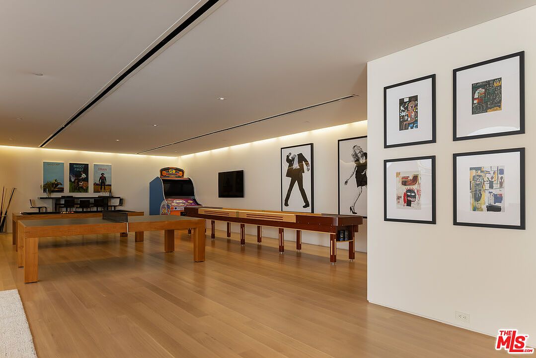 This is a spacious basement featuring a ping pong table, shuffleboard table, and an arcade game, suggesting a recreational area. The walls are adorned with framed artwork, and the hardwood flooring adds warmth to the space. The room is well-lit, creating an inviting atmosphere for entertainment.