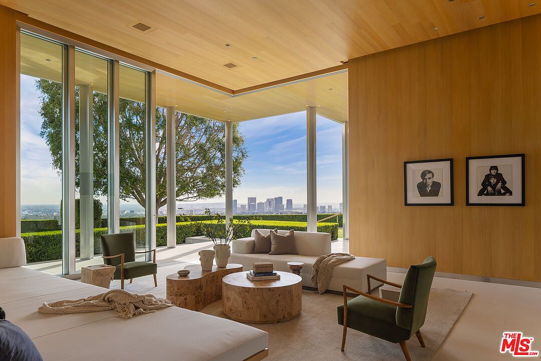 This is a bright and airy living room featuring floor-to-ceiling windows that offer panoramic views of the city skyline and lush greenery. The room is furnished with modern, minimalist furniture, including a large sectional sofa, unique wooden coffee tables, and stylish armchairs. The light wood paneling on the walls and ceiling adds warmth and sophistication to the space.