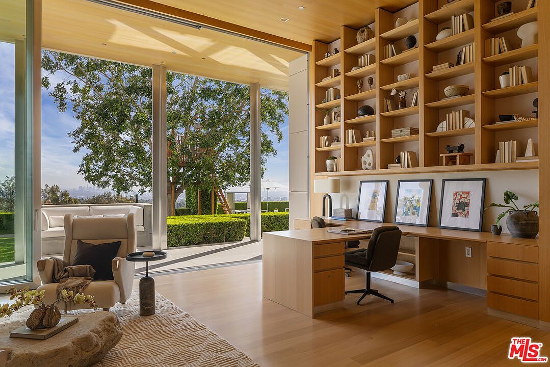 This is an interior shot of a modern office space featuring a built-in wooden bookshelf and desk. The room has large glass doors that open to an outdoor area with lush greenery and a glimpse of the city skyline. The space is well-lit and appears to be designed for both functionality and relaxation.