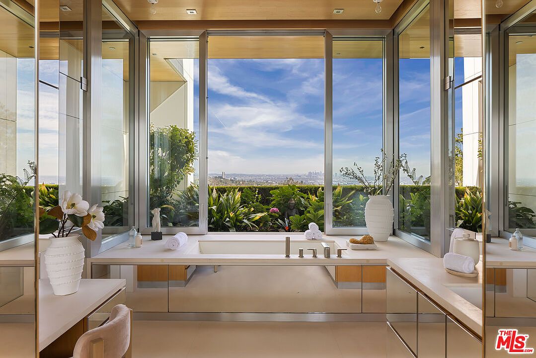 This is a luxurious primary bathroom featuring a large soaking tub positioned in front of a wall of windows that offer a stunning view of lush greenery and the city skyline. The bathroom is designed with a modern aesthetic, incorporating clean lines, light-colored countertops, and mirrored accents that enhance the sense of space and light. Vases with flowers and decorative items add a touch of elegance.