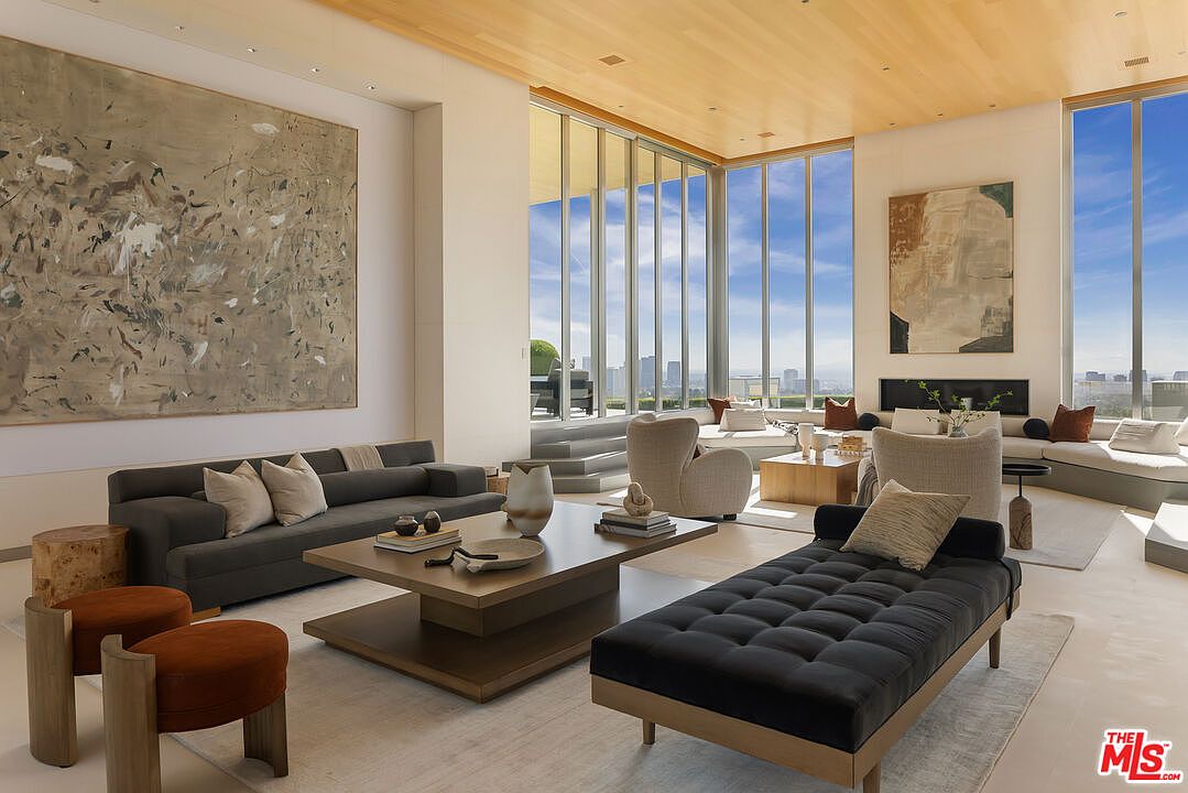 This is a spacious and luxurious living room featuring floor-to-ceiling windows with city views. The room is furnished with a gray sofa, a black chaise lounge, and a unique coffee table, complemented by abstract art pieces. The overall design is modern and sophisticated, creating an inviting and elegant atmosphere.