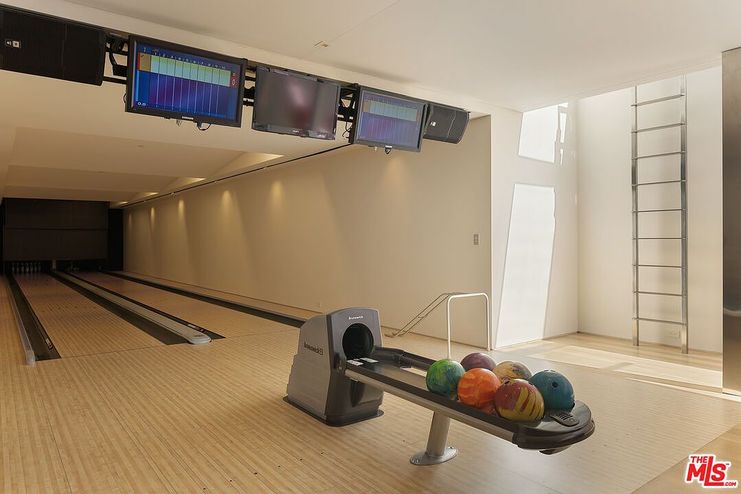 This interior shot showcases a private bowling alley, complete with two lanes, overhead scoring monitors, and a ball return system holding several bowling balls. The space features light wood flooring, neutral-toned walls, and a modern ladder leading to an upper level, creating a unique and luxurious recreational area. The perspective is from the approach area, looking towards the pins.