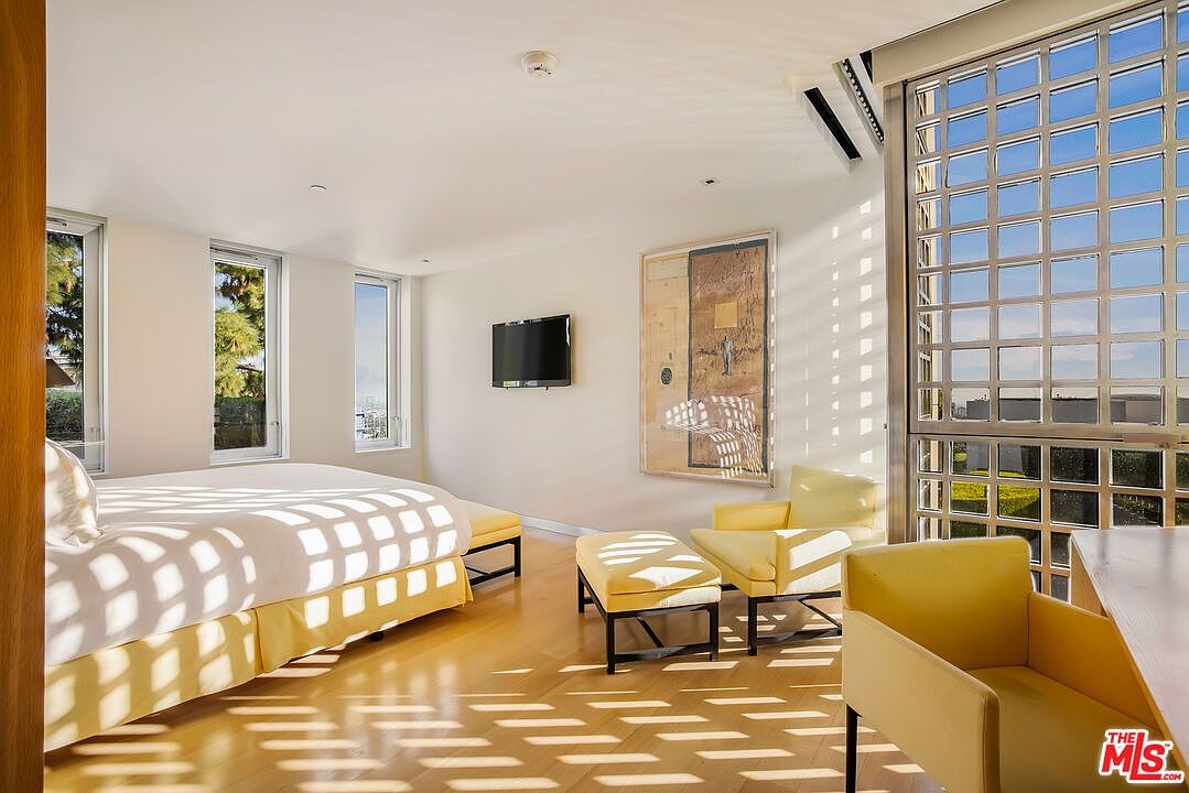 This is a bright and airy primary bedroom featuring a large bed with yellow accents, a seating area with yellow chairs and ottoman, and a modern art piece on the wall. The room is illuminated by natural light streaming through large windows, creating a warm and inviting atmosphere. The hardwood floors and minimalist design contribute to a clean and contemporary aesthetic.
