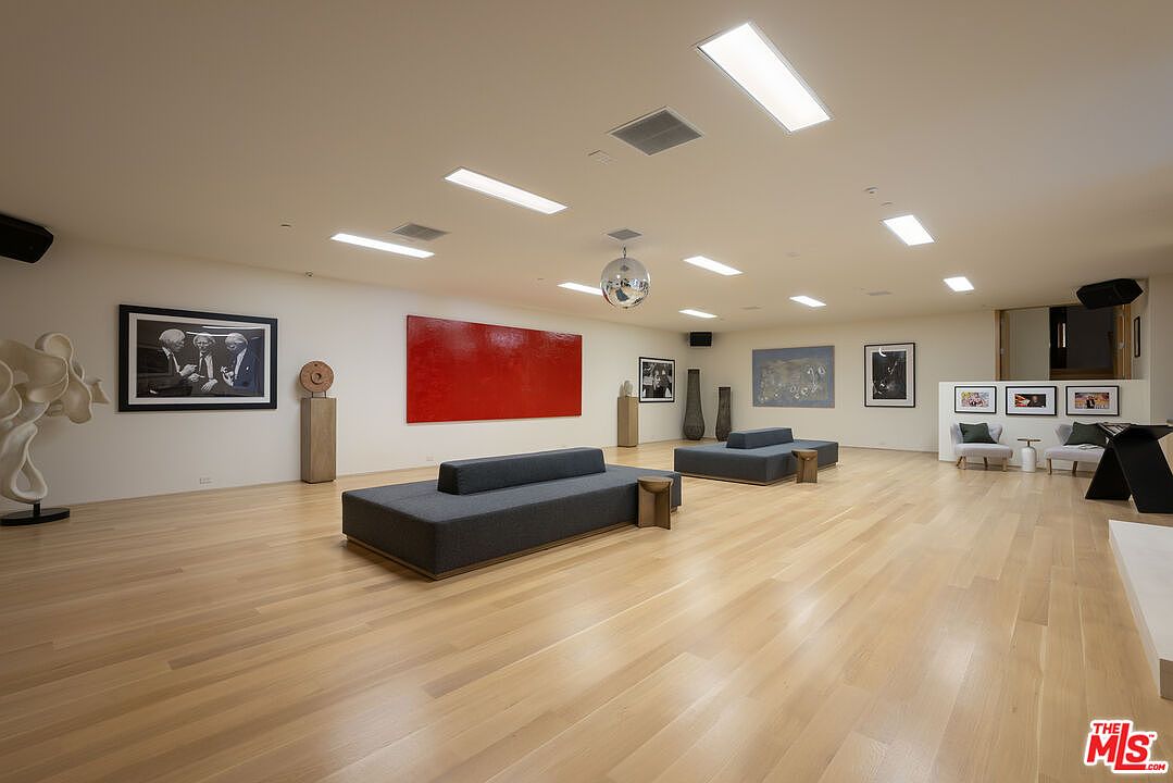 This expansive living room showcases a modern art gallery aesthetic with light wood flooring and neutral walls. The room features contemporary sofas, various art pieces including a large red canvas and framed photographs, and a disco ball hanging from the ceiling. Recessed lighting and rectangular ceiling lights illuminate the space, creating a bright and airy atmosphere.