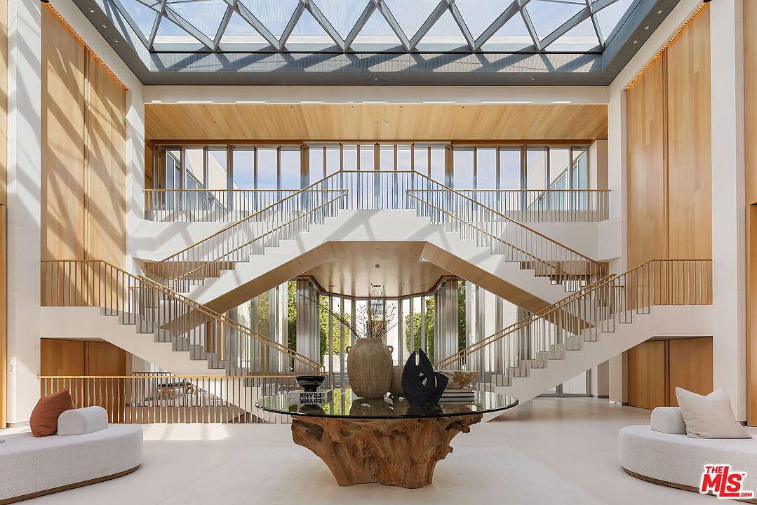 This grand foyer features a symmetrical staircase design with light wood accents and white risers, leading to an upper level with large windows. A unique, organically shaped table with a glass top serves as a focal point, complemented by modern seating. The space is bathed in natural light from a large skylight, creating an open and airy atmosphere.