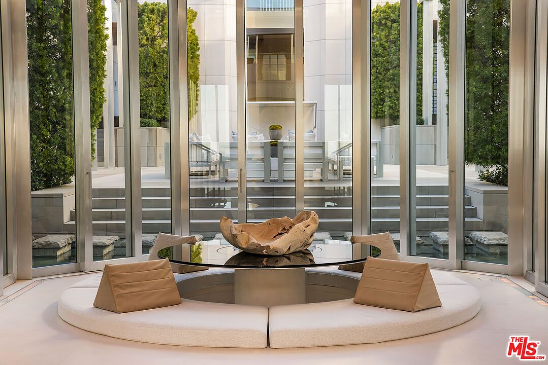 This sunroom features a unique circular seating area with a glass-topped table and sculptural centerpiece. Large glass doors and windows provide ample natural light and views of the exterior garden and architectural details. The space exudes a modern and serene ambiance, perfect for relaxation and contemplation.