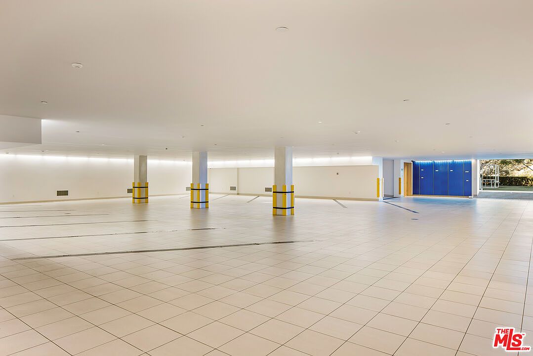 The image showcases a large, open basement space with tiled flooring and white walls. Support columns with yellow and black safety bands are visible, and there are blue lockers in the background. The space appears clean and well-lit, suggesting potential for various uses such as a recreation area or storage.