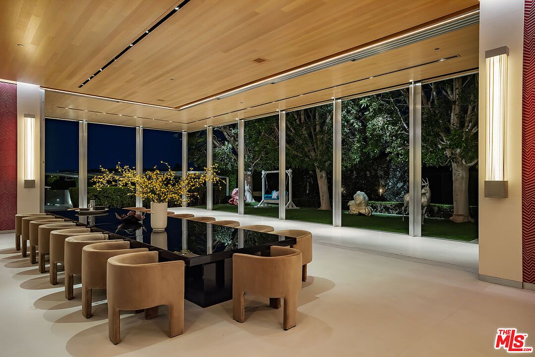 This is a spacious dining room featuring a long, dark-colored dining table surrounded by numerous beige chairs. Large windows offer a view of the outdoor garden, complete with sculptures and a swing. The room is well-lit with modern wall sconces and recessed lighting, creating an elegant and inviting atmosphere.