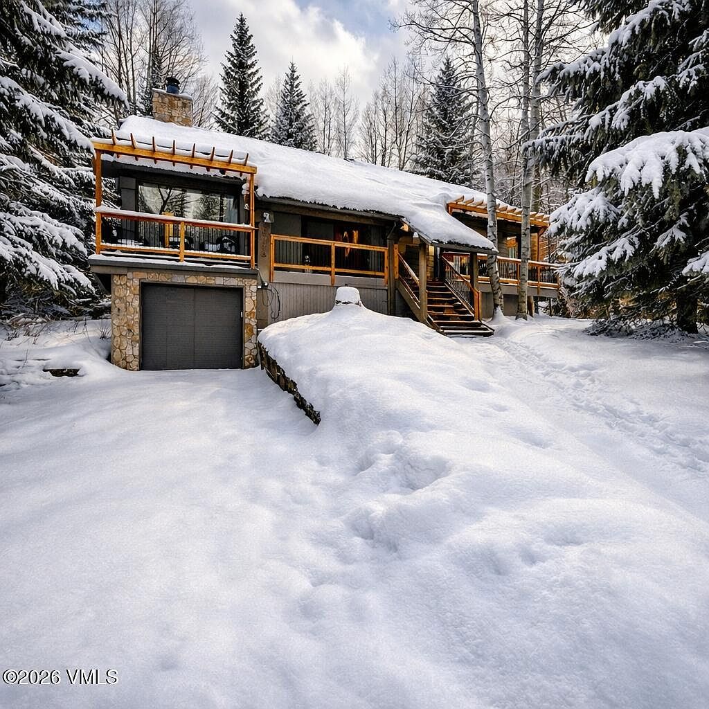 This charming mountain home is nestled in a snowy, wooded landscape, featuring a rustic stone and wood exterior with a prominent garage and elevated deck. The house is framed by tall pine and aspen trees, creating a serene and secluded winter retreat atmosphere. A set of wooden stairs leads up to the main entrance, highlighting the home's cozy, elevated design.