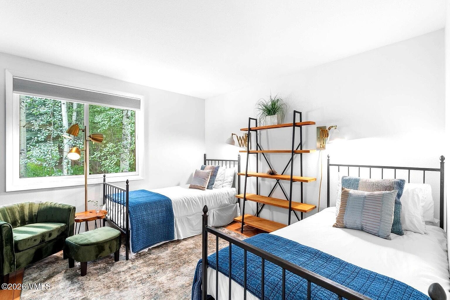 This bright and inviting guest bedroom features two twin beds with black metal frames, dressed in crisp white linens and vibrant blue quilted coverlets. A tall, open-concept wooden shelving unit stands between the beds, while a plush green velvet armchair and matching ottoman provide a cozy seating area near the window. The room is bathed in natural light, creating a serene and comfortable atmosphere perfect for guests.