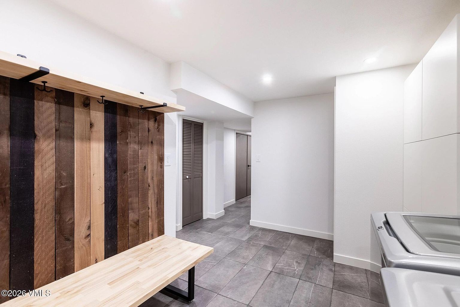 This functional laundry room features a modern aesthetic with a striking accent wall made of reclaimed wood planks, complete with built-in hooks and a matching wooden bench. The space includes sleek white cabinetry and gray tile flooring, leading into a hallway with louvered doors. The perspective is a wide-angle shot that highlights the clean, organized layout and the blend of rustic and contemporary design elements.