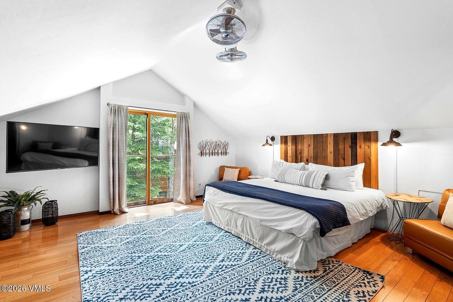 This spacious primary bedroom features a vaulted ceiling, a large bed with a rustic wood headboard, and a cozy seating area. A sliding glass door leads to a private balcony, allowing natural light to fill the room, while a large wall-mounted television and a patterned area rug add comfort and style. The room exudes a warm, inviting atmosphere with its blend of modern amenities and natural wood accents.
