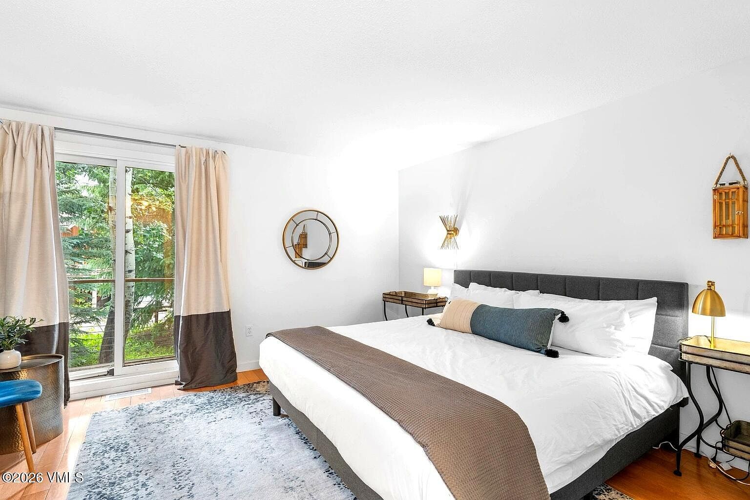 This bright and airy primary bedroom features a large bed with a dark grey upholstered headboard, dressed in crisp white linens and a brown runner. A sliding glass door provides ample natural light and a view of the surrounding trees, while a stylish area rug adds texture to the hardwood flooring. The room is accented with modern wall sconces, a circular mirror, and elegant bedside tables, creating a serene and inviting atmosphere.