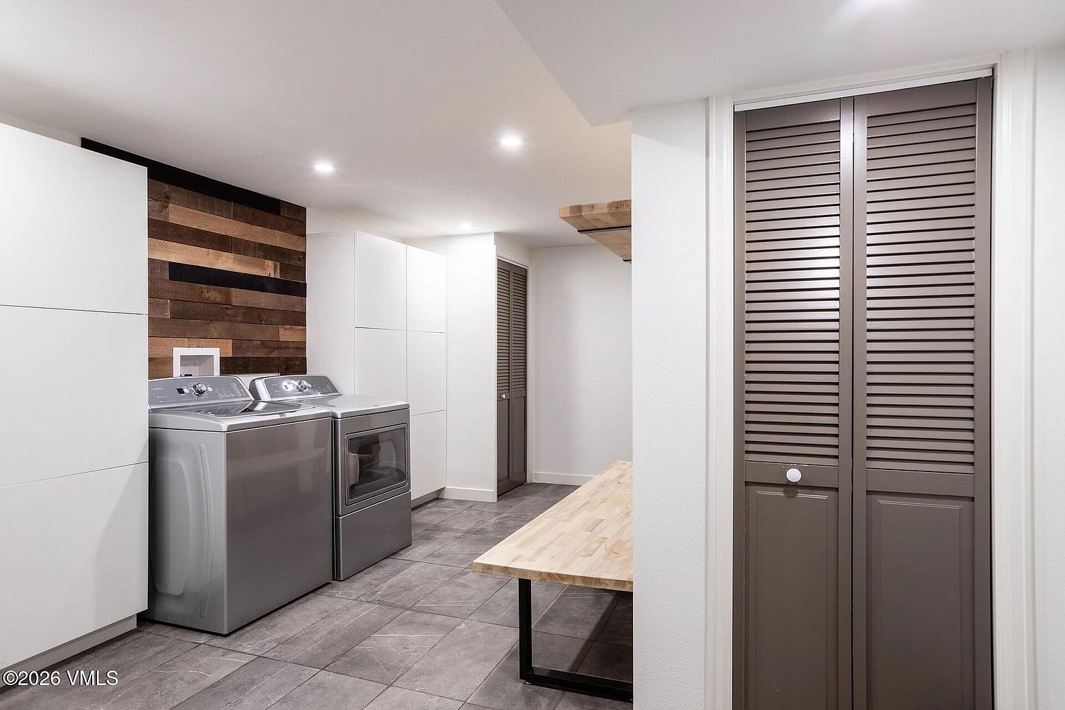 This modern laundry room features sleek stainless steel appliances set against a striking wood-plank accent wall. A long, light-wood folding table provides ample workspace, while dark grey floor tiles and louvered closet doors add a sophisticated, contemporary touch. The space is brightly lit with recessed ceiling lights, creating a clean and functional environment.