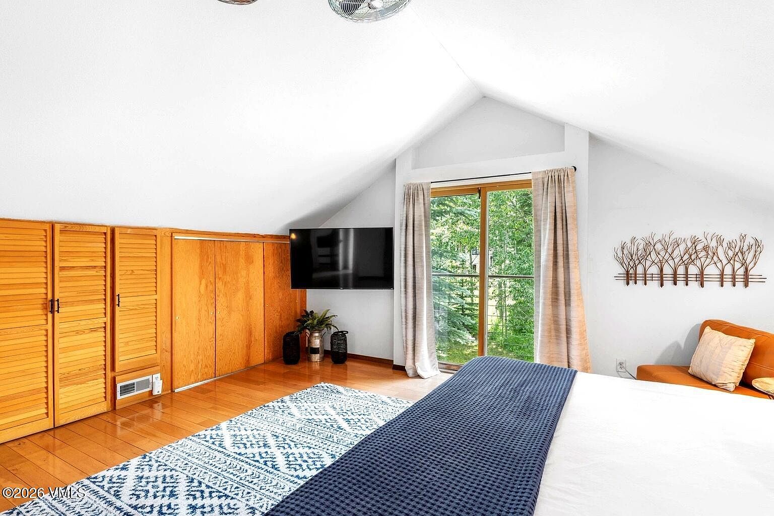 This primary bedroom features a vaulted ceiling and warm wood flooring, creating a cozy and inviting atmosphere. A large bed with a blue textured throw sits in the foreground, while a wall-mounted television and a sliding glass door leading to a balcony provide functional and scenic elements. The room is accented by built-in wooden cabinetry and a decorative tree-themed wall art piece, blending rustic charm with modern comfort.