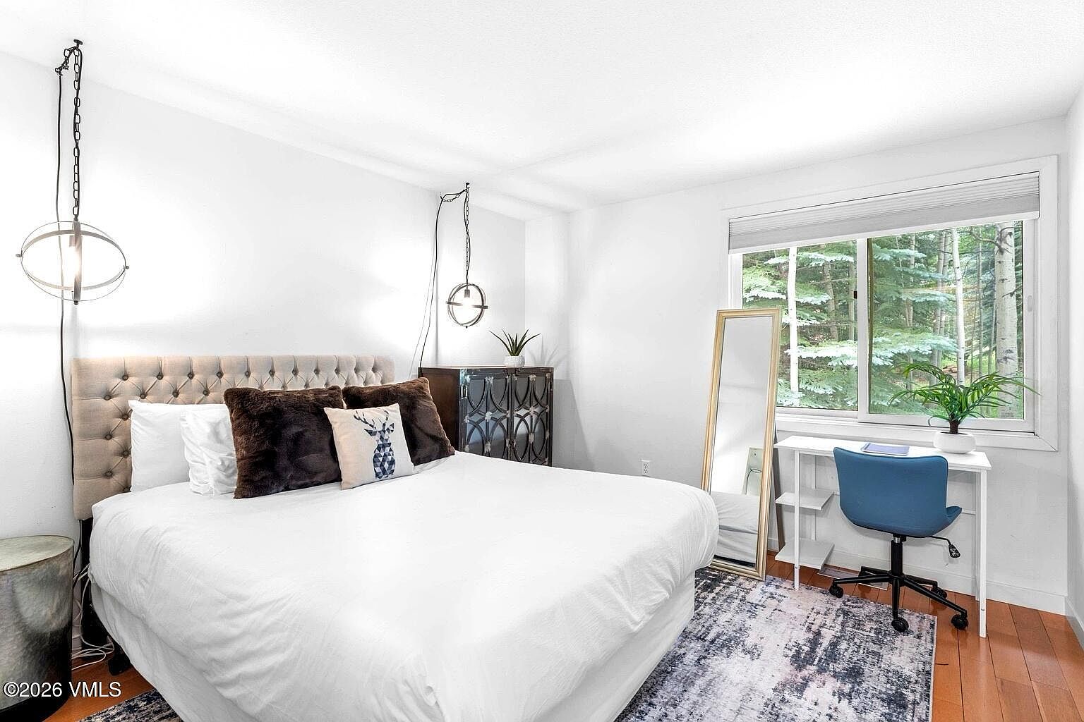 This bright and modern bedroom features a plush, tufted headboard and a crisp white bedspread, accented by dark brown decorative pillows. A dedicated workspace with a blue ergonomic chair is positioned by a large window that offers a serene view of the surrounding forest. The room is styled with contemporary lighting fixtures and a full-length floor mirror, creating a clean and inviting atmosphere.