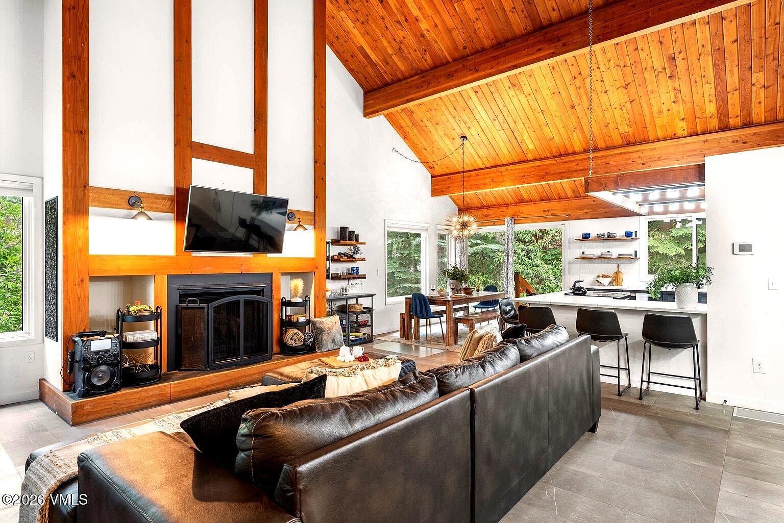 This open-concept living space features a striking floor-to-ceiling fireplace with wood accents and a vaulted, wood-paneled ceiling that adds warmth and character. The room flows seamlessly into a dining area and kitchen, creating an inviting atmosphere perfect for entertaining. Large windows and glass doors provide abundant natural light and views of the surrounding greenery, enhancing the home's airy, modern mountain aesthetic.