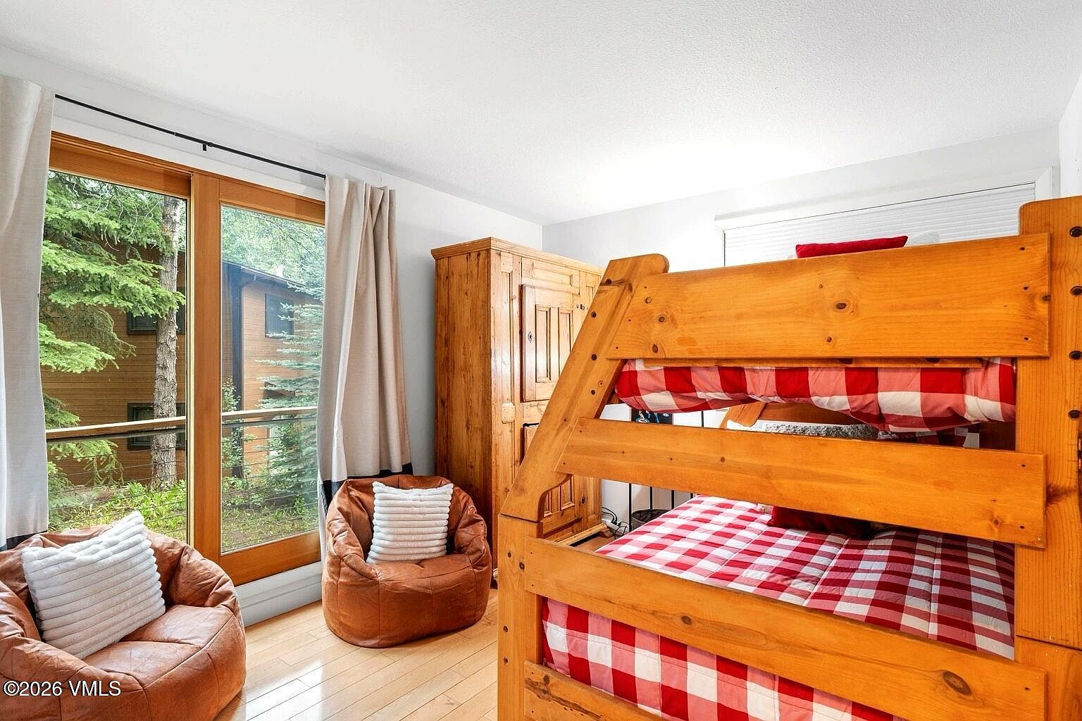 This cozy bedroom features a rustic wooden bunk bed dressed in red and white checkered bedding, complemented by two matching leather beanbag chairs. A large wooden armoire provides storage, while a floor-to-ceiling glass door offers a view of the surrounding forest, filling the room with natural light. The space has a warm, cabin-like aesthetic perfect for a mountain retreat.