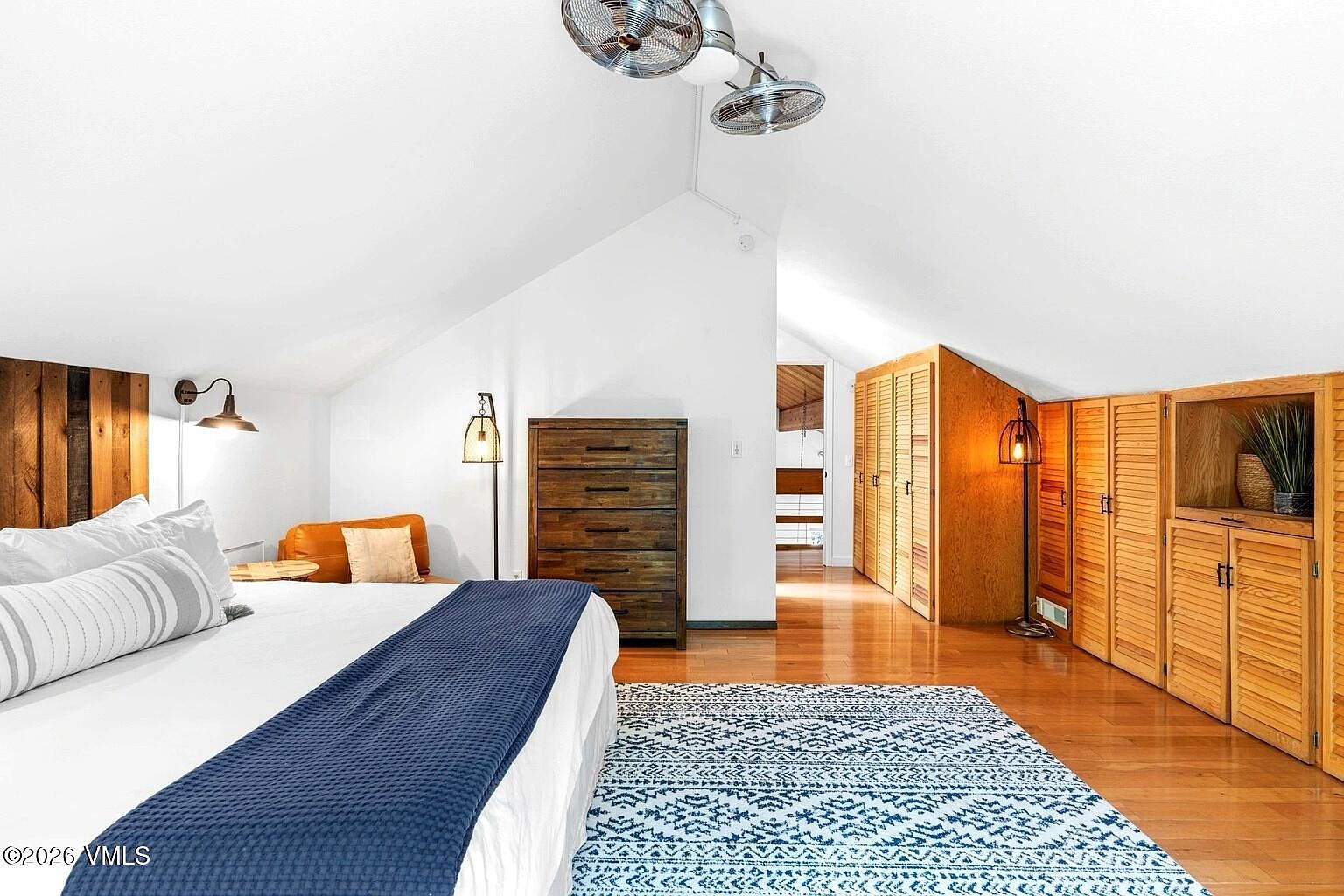 This spacious attic bedroom features a vaulted ceiling with dual ceiling fans and warm hardwood flooring. The room is furnished with a large bed topped with a navy blue throw, a rustic wooden dresser, and built-in louvered closet doors that provide ample storage. A patterned area rug adds texture to the space, while a small seating area near the window creates a cozy, inviting atmosphere.