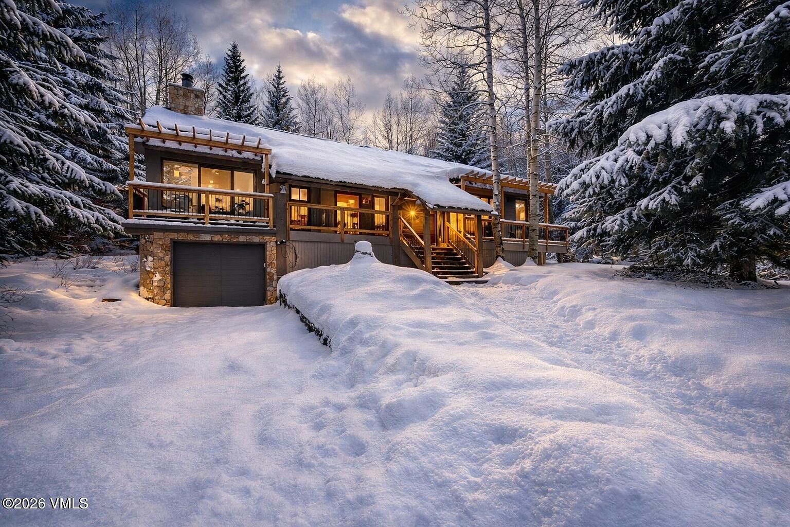 This charming mountain home is captured in a picturesque winter setting, blanketed in deep, fresh snow. The structure features a rustic aesthetic with wood siding, a stone-accented garage base, and a welcoming elevated deck with a pergola-style roof. Warm interior lighting glows through the windows, creating a cozy and inviting contrast against the cool, twilight sky and surrounding snow-covered evergreen trees.