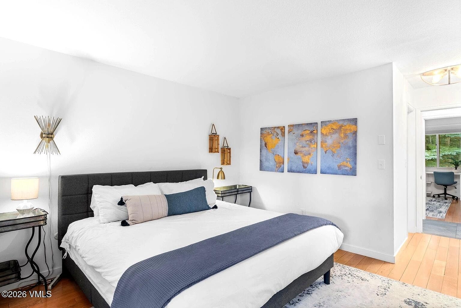 This bright and airy primary bedroom features a modern aesthetic with a plush, dark gray upholstered bed frame as the centerpiece. The room is accented by a triptych world map art piece, decorative wall lanterns, and a soft blue runner, creating a serene and inviting atmosphere. A doorway in the background provides a glimpse into a well-lit home office, enhancing the sense of flow and functionality within the home.