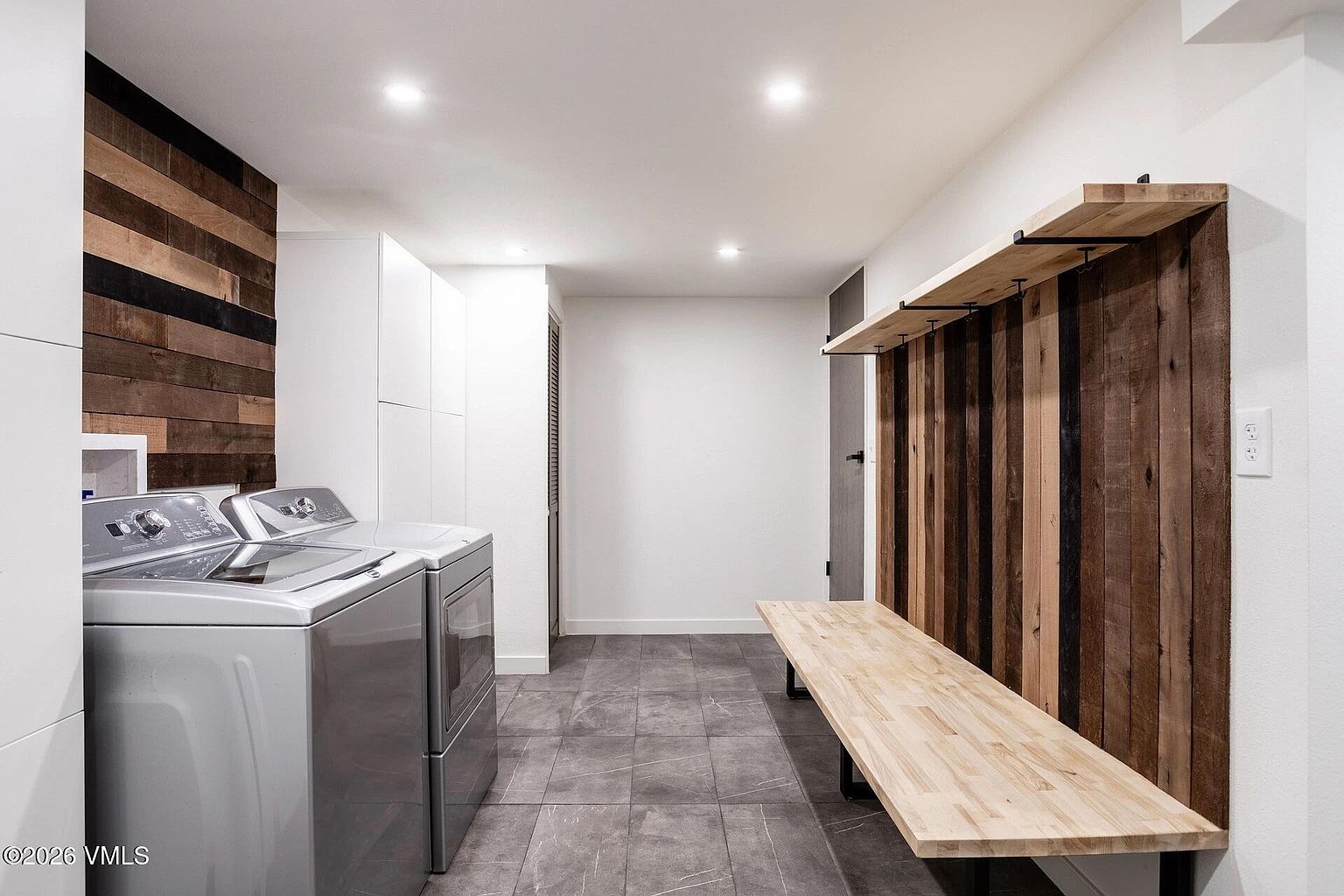 This modern laundry room features a sleek, industrial-inspired design with a combination of light wood accents and dark, rustic wood paneling. The space includes a high-efficiency washer and dryer set, a built-in wooden bench with overhead shelving, and dark grey tile flooring. The clean lines and recessed lighting create a functional yet stylish utility area.