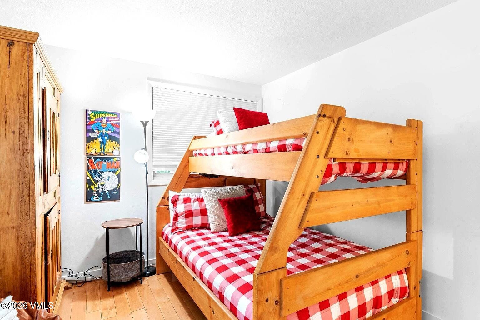 This cozy bedroom features a sturdy wooden bunk bed dressed in red and white checkered bedding, creating a playful and inviting atmosphere. A small side table with a floor lamp sits next to the bed, while comic book-themed wall art adds a touch of personality to the space. The room is finished with light-toned wood flooring and neutral walls, making it a functional and charming space for guests or children.
