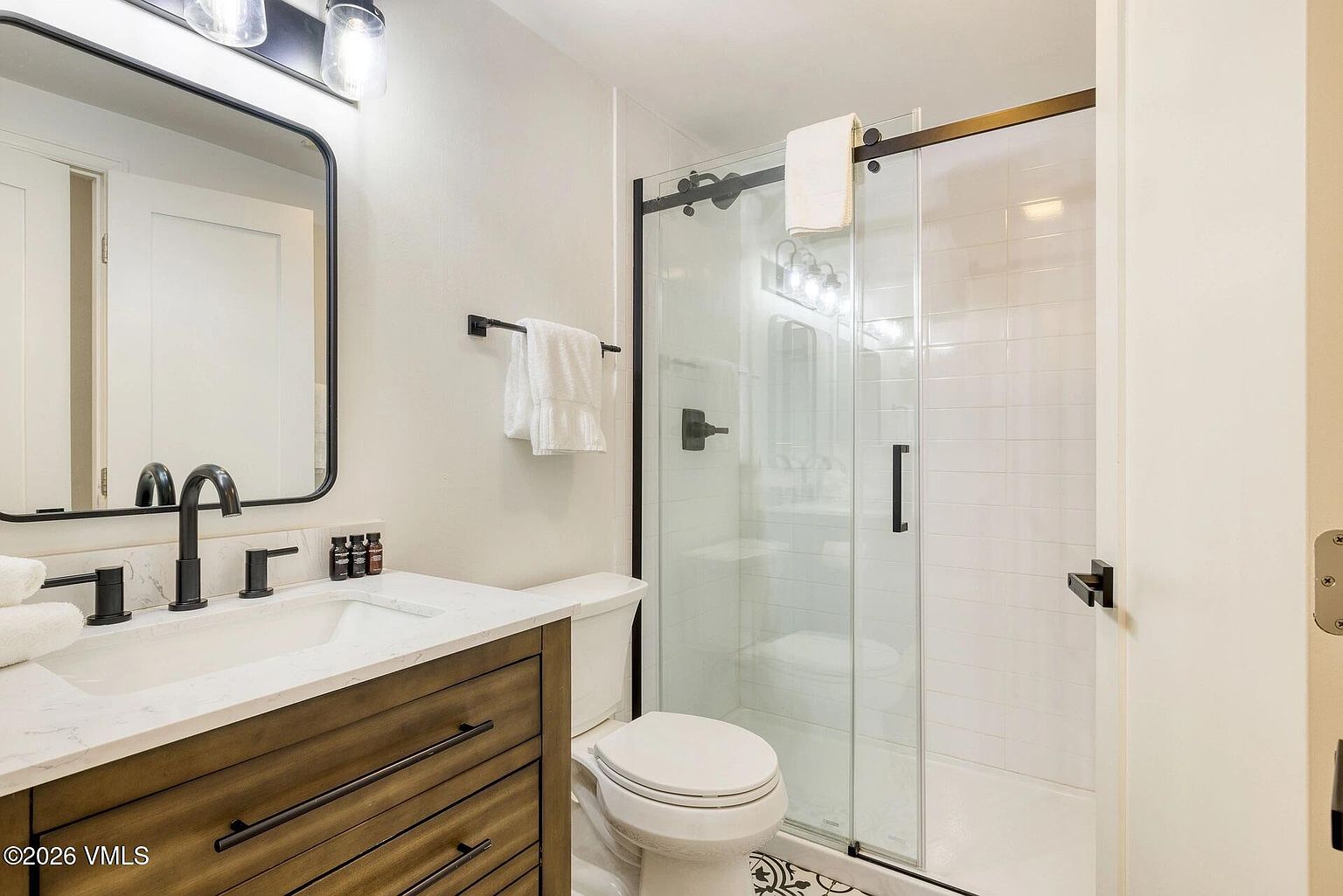 This modern bathroom features a sleek vanity with a white countertop, a dark-framed mirror, and matte black fixtures. A glass-enclosed walk-in shower with white subway tile adds a clean, contemporary aesthetic to the space. The room is well-lit and compact, showcasing a functional and stylish design suitable for a guest or secondary bathroom.