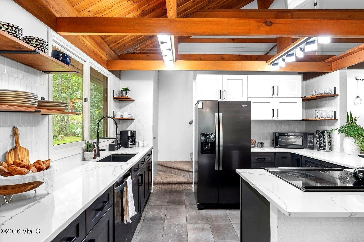 This modern kitchen features a striking contrast between dark lower cabinetry and crisp white upper cabinets, complemented by white quartz countertops and a subway tile backsplash. The space is defined by its vaulted wood-beamed ceiling, which adds warmth and architectural character, while a large black refrigerator and integrated appliances provide a sleek, functional aesthetic. The perspective is a wide-angle shot from the kitchen island, showcasing the open layout that leads toward a carpeted staircase in the background.