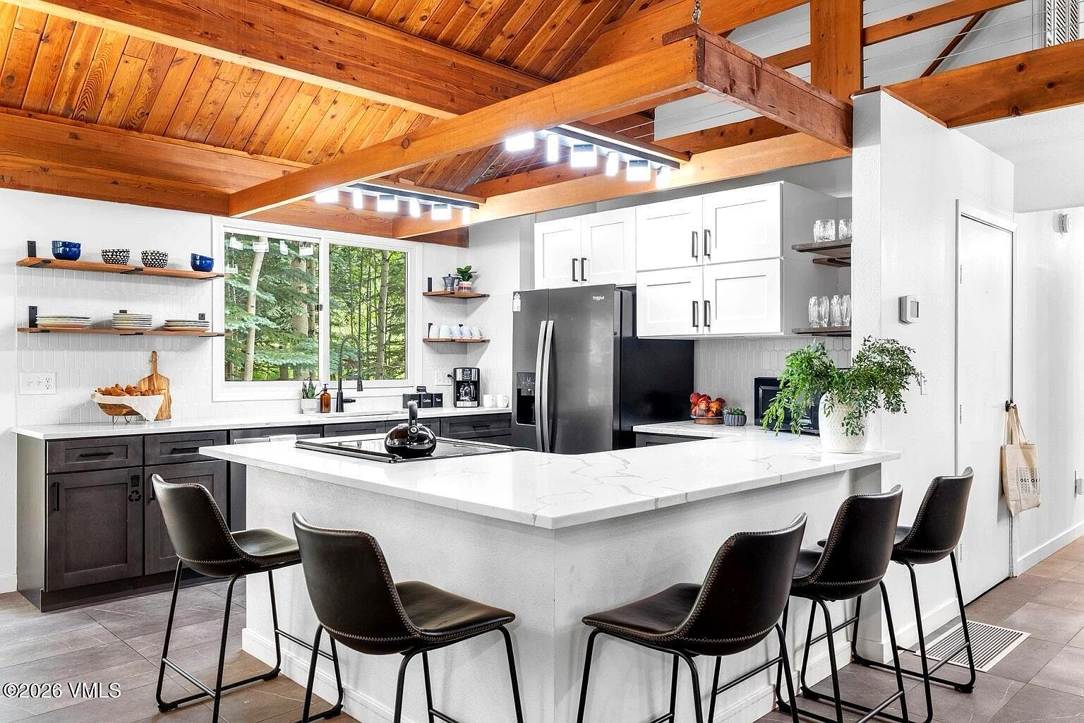This modern, open-concept kitchen features a large white quartz island with bar seating, dark lower cabinetry, and crisp white upper cabinets. The space is highlighted by a striking vaulted wood-paneled ceiling with exposed beams and a contemporary linear light fixture. A large window above the sink provides a serene view of the surrounding forest, creating a bright and inviting atmosphere.