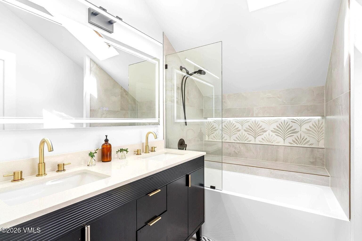 This modern primary bathroom features a sleek black double vanity with gold hardware and a white countertop, complemented by a large, illuminated mirror. A glass-enclosed shower and tub combination is accented with decorative leaf-patterned tile work and integrated lighting. The space is bright and contemporary, showcasing a clean, high-end design aesthetic.