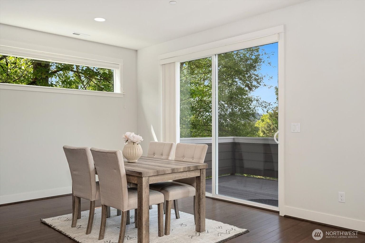 This bright dining area features a light wood-toned table surrounded by four upholstered chairs, set atop a textured area rug. A large sliding glass door provides access to an outdoor deck, while a horizontal window above the table adds extra natural light to the space. The room is finished with dark wood flooring and neutral walls, creating a clean and inviting atmosphere.