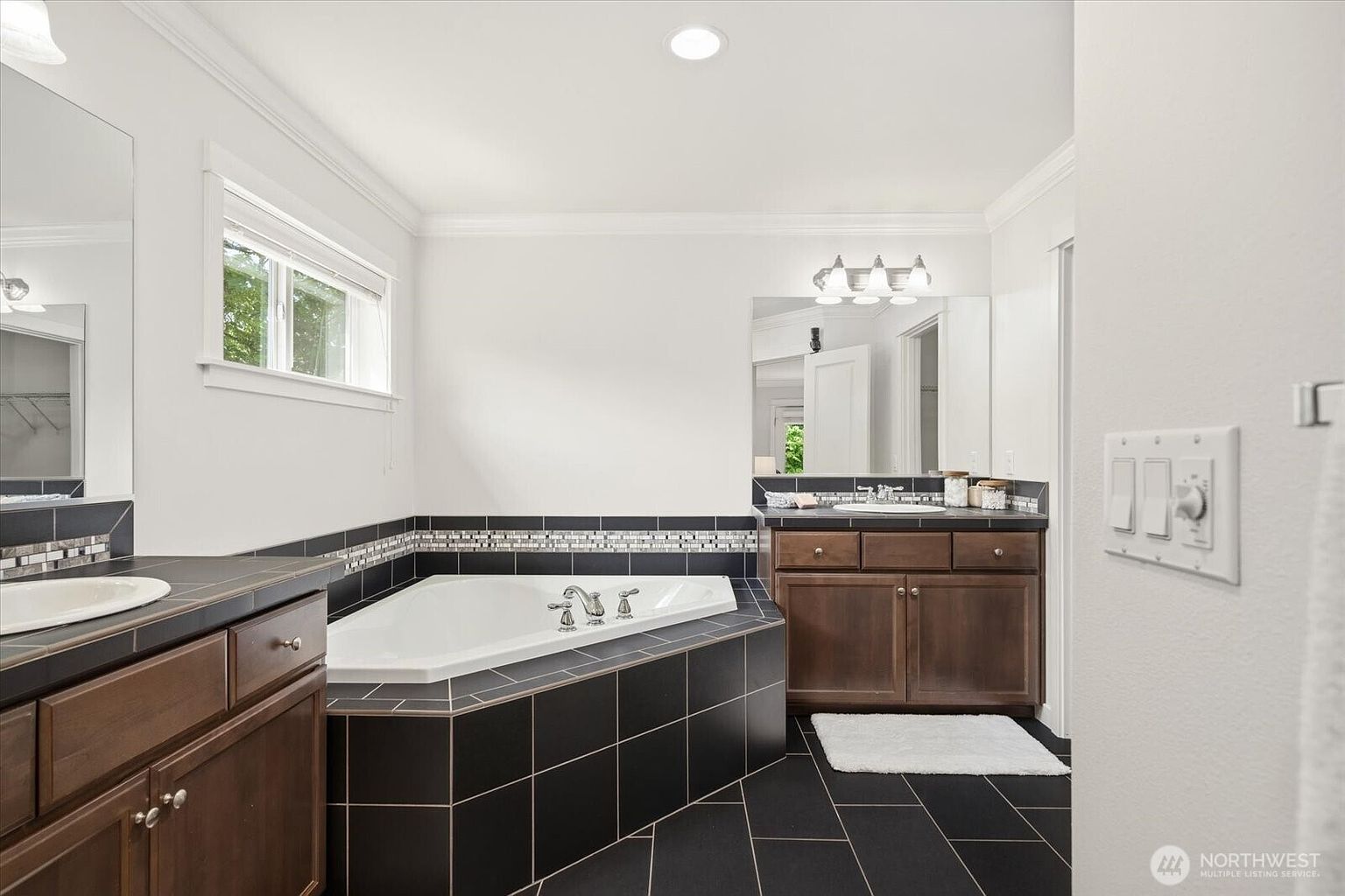 This primary bathroom features a corner soaking tub surrounded by dark, square-tiled decking and a decorative mosaic accent border. Dual vanities with dark wood cabinetry and light-colored countertops are positioned on either side of the tub, each complemented by a large wall-mounted mirror and overhead lighting. The space is finished with matching dark floor tiles, creating a cohesive and traditional aesthetic.