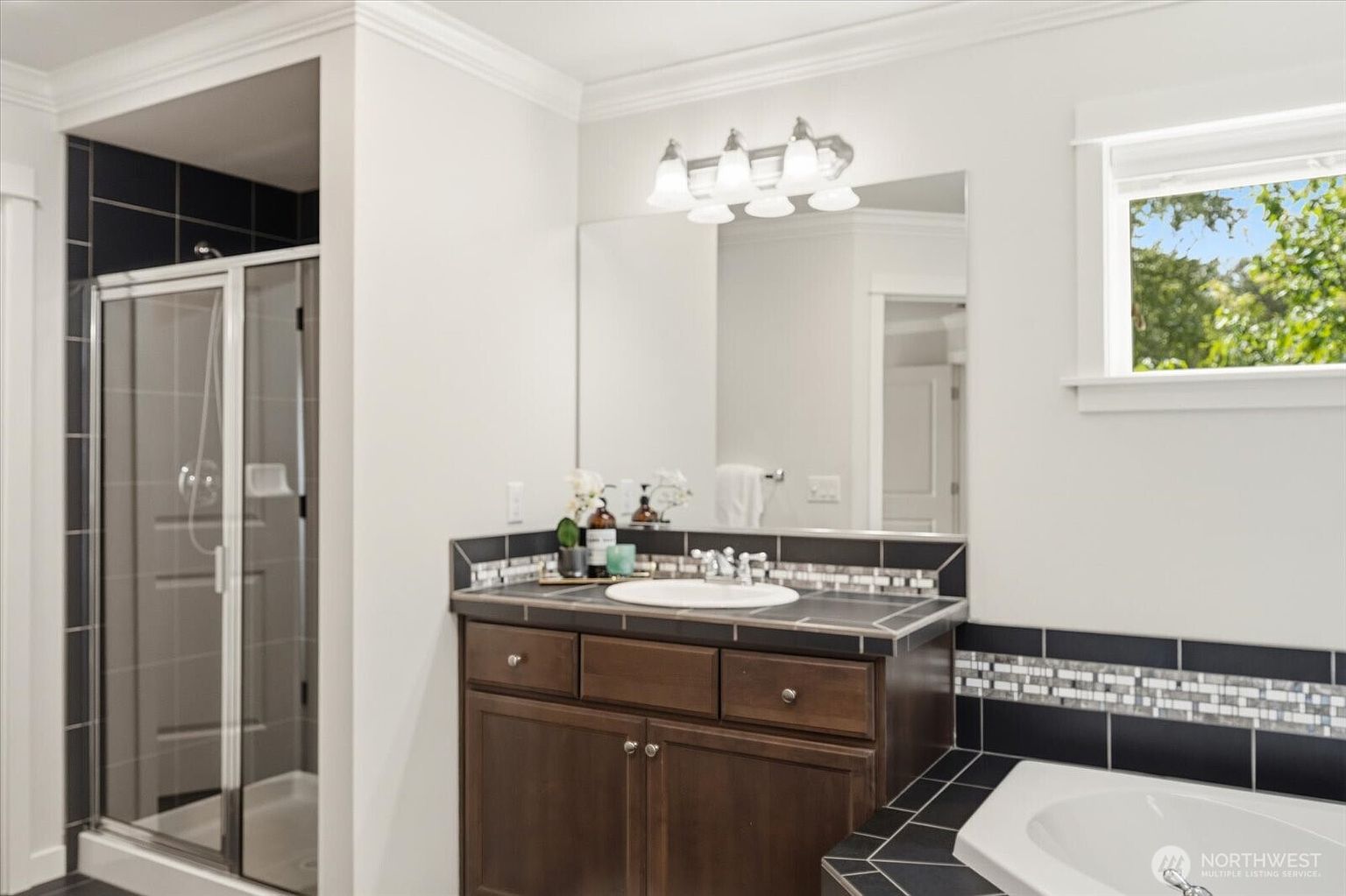 This bathroom features a dark wood vanity with a light-colored countertop and an integrated sink, complemented by a large, frameless mirror and a multi-light fixture above. To the left, a glass-enclosed shower is framed by dark, grid-patterned wall tiles that contrast with the crisp white walls. A corner soaking tub is partially visible in the foreground, and a window provides natural light to the space.