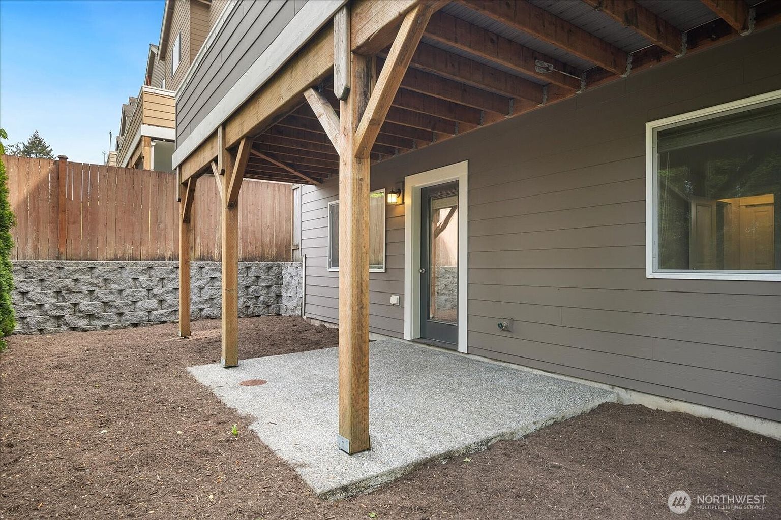 This image captures a ground-level concrete patio situated beneath an elevated wooden deck. The space features a glass-paneled door providing access to the home's interior, with a retaining wall made of stacked stone blocks bordering the adjacent yard area. The perspective is a low-angle shot looking toward the house, highlighting the structural wooden support posts and the transition between the paved patio and the surrounding dirt yard.