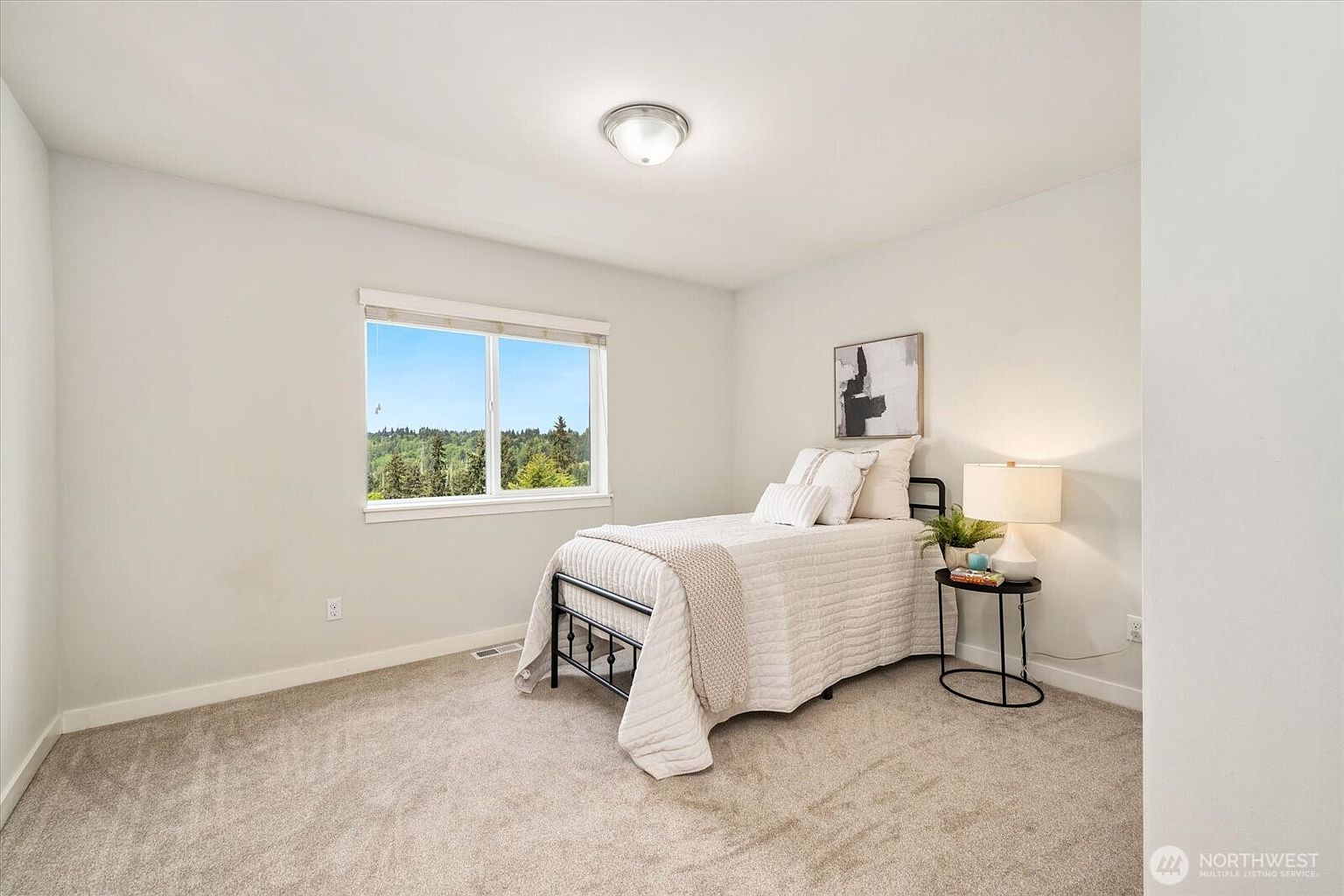 This bright and neutral guest bedroom features plush carpeting and a single bed dressed in white linens. A window on the left wall provides natural light and a view of the surrounding trees, while a small circular side table with a lamp sits beside the bed. The room is decorated with a simple piece of wall art, creating a clean and inviting atmosphere.