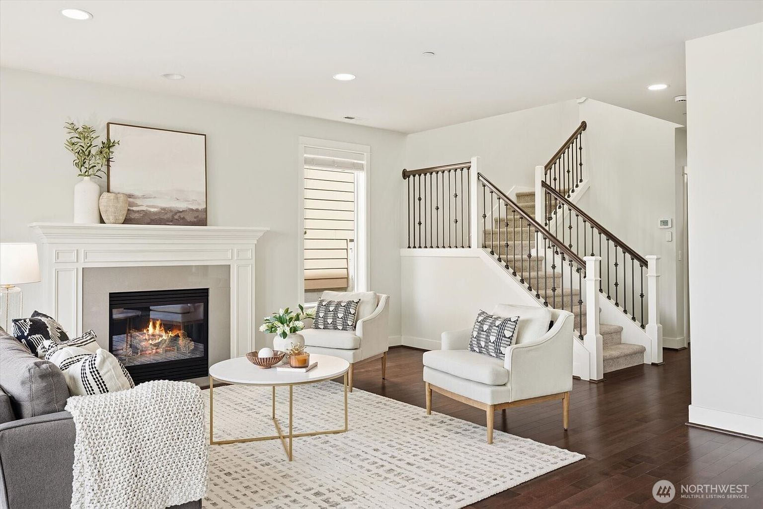 This bright and inviting living room features a cozy fireplace with a white mantel and a neutral-toned surround as its focal point. The space is furnished with a comfortable gray sofa and two matching armchairs arranged around a circular coffee table on a textured area rug. A staircase with dark wood railings and white balusters adds architectural interest, while the dark hardwood floors and recessed lighting create a warm, polished atmosphere.