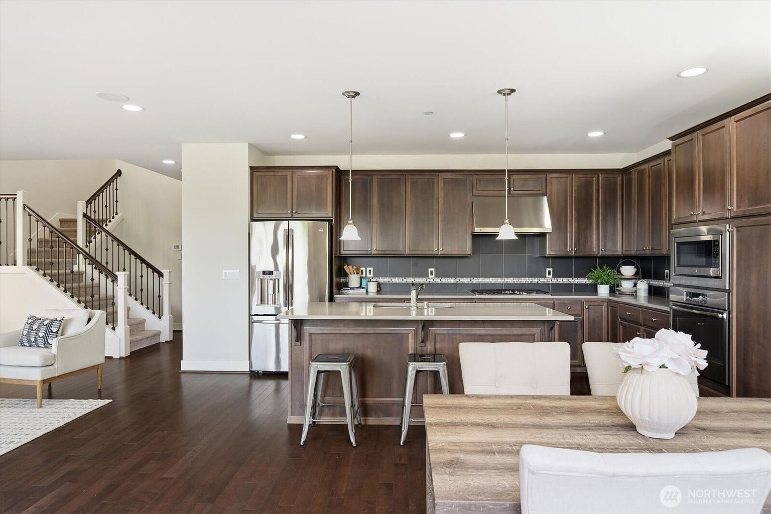 This open-concept kitchen features dark wood cabinetry, a central island with bar seating, and stainless steel appliances. The space is illuminated by pendant lighting over the island and recessed ceiling lights, with a staircase visible in the background. The warm wood flooring and neutral color palette create a cohesive and inviting atmosphere.