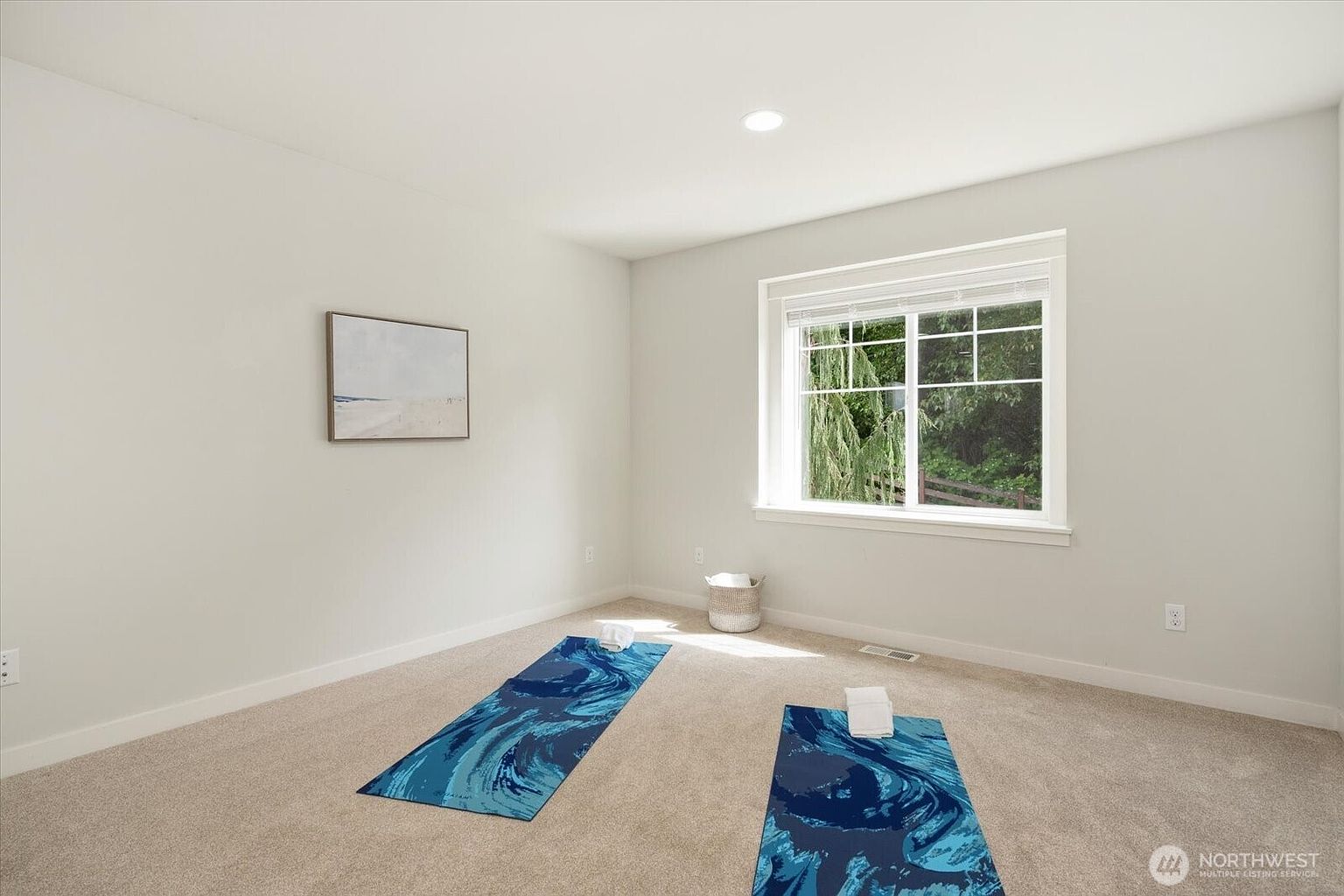 This bright and airy room is currently staged as a home yoga or fitness space, featuring neutral carpeting and light-colored walls. Two blue patterned yoga mats are laid out on the floor, each with a small white towel, while a large window provides natural light and a view of the greenery outside. A piece of abstract wall art and a woven basket in the corner add a touch of minimalist decor to the clean, open space.