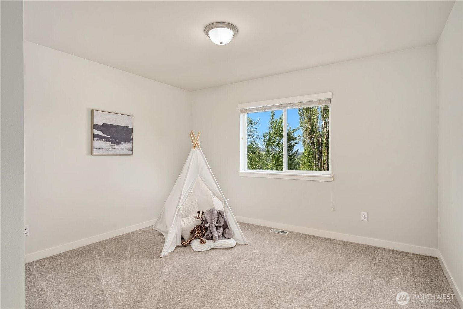 This bright and neutral bedroom features soft carpeting and a minimalist aesthetic, currently staged as a child's play area with a white teepee tent. A large window allows for ample natural light and provides a view of the surrounding trees, while a simple framed piece of art hangs on the wall. The room is finished with a standard ceiling light fixture and clean, white-painted walls, offering a versatile space for various uses.