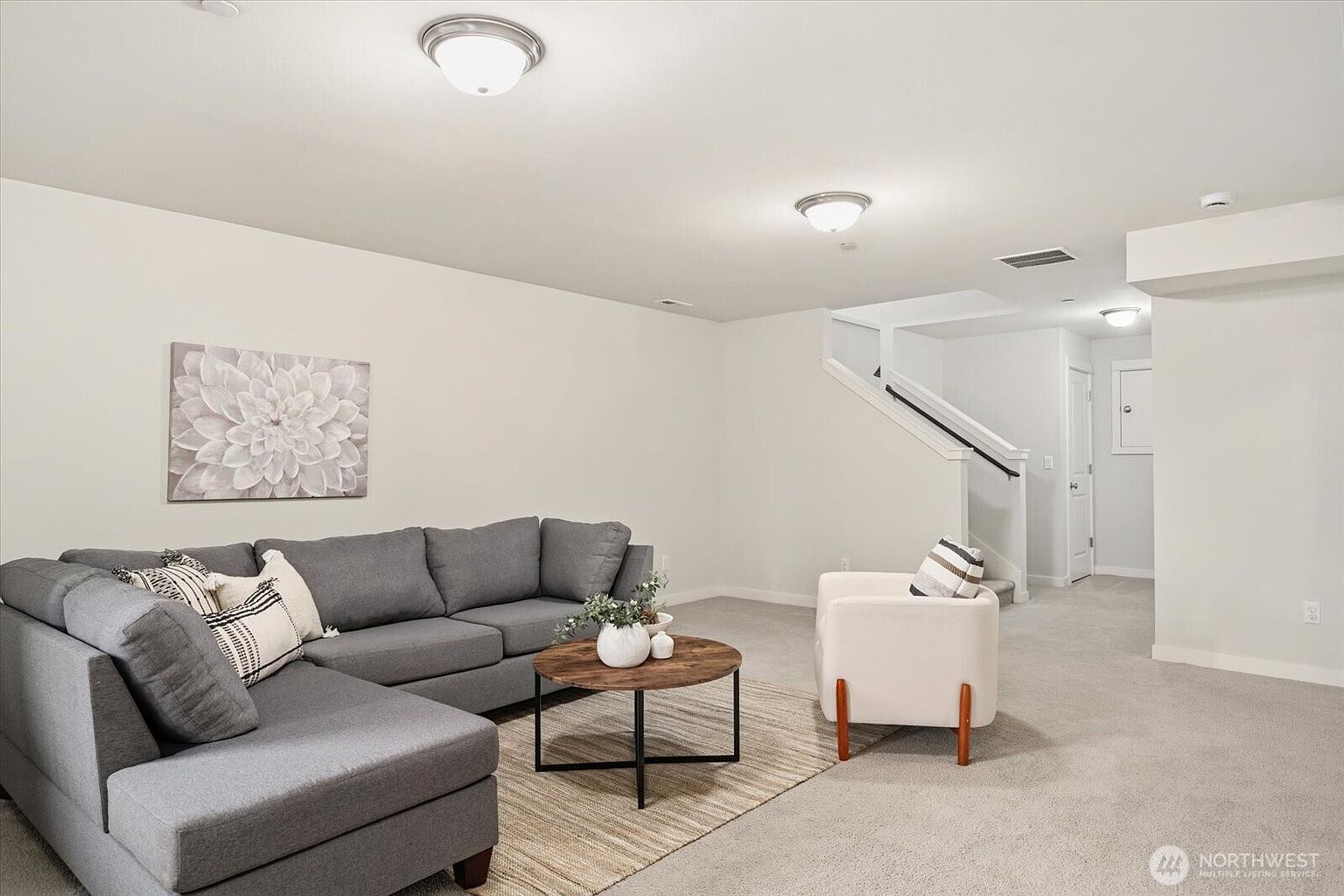 This spacious, neutrally-toned living room features a large grey sectional sofa arranged on a textured area rug, complemented by a round wooden coffee table. A modern white armchair sits to the right, while a staircase with a dark handrail leads to an upper level in the background. The room is illuminated by two flush-mount ceiling lights, creating a bright and inviting atmosphere suitable for relaxation.