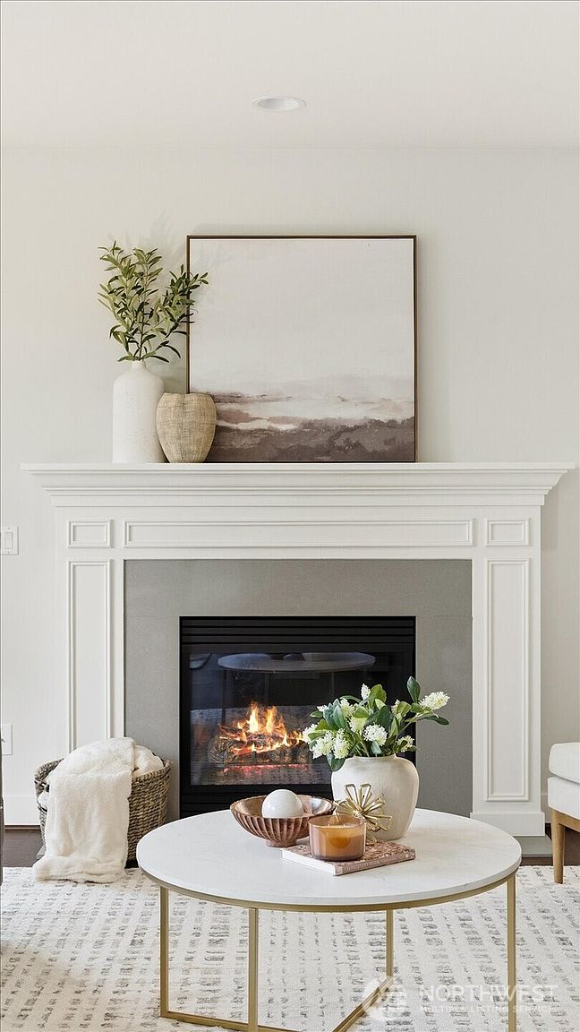 This living room features a central fireplace with a white mantel and a neutral-toned surround, serving as the focal point of the space. A round coffee table with a light-colored top sits in the foreground, adorned with decorative items, while a textured rug and a basket with a throw blanket add warmth to the room. The aesthetic is clean and modern, characterized by a soft, neutral color palette and tasteful decor.