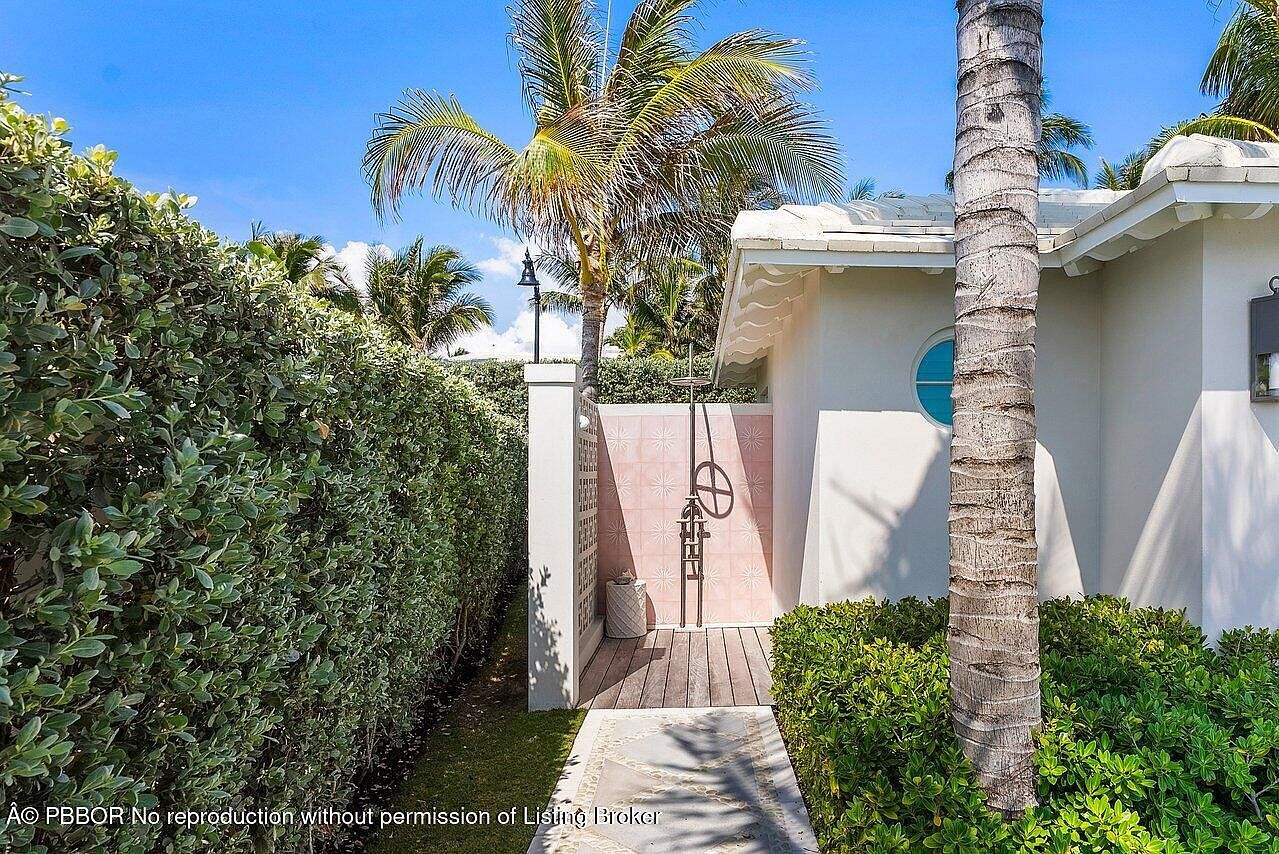 This image showcases a charming outdoor shower area, nestled within lush greenery and featuring a decorative pink tiled wall. The shower is accessible via a stone pathway and wooden deck, adding to the secluded and luxurious feel. A palm tree and the side of a white building are also visible, enhancing the tropical ambiance.