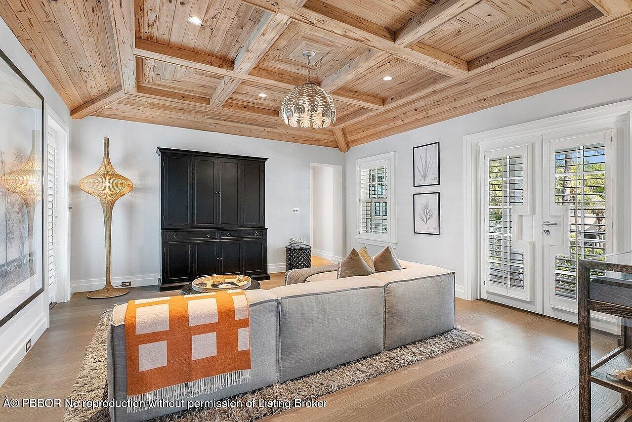 This living room features a striking wood-paneled ceiling with recessed lighting and a decorative chandelier. A large, comfortable sectional sofa sits on a textured rug, complemented by a dark wood cabinet and a unique floor lamp. The room is bright and airy, with natural light streaming in through shuttered windows and doors, creating a warm and inviting atmosphere.