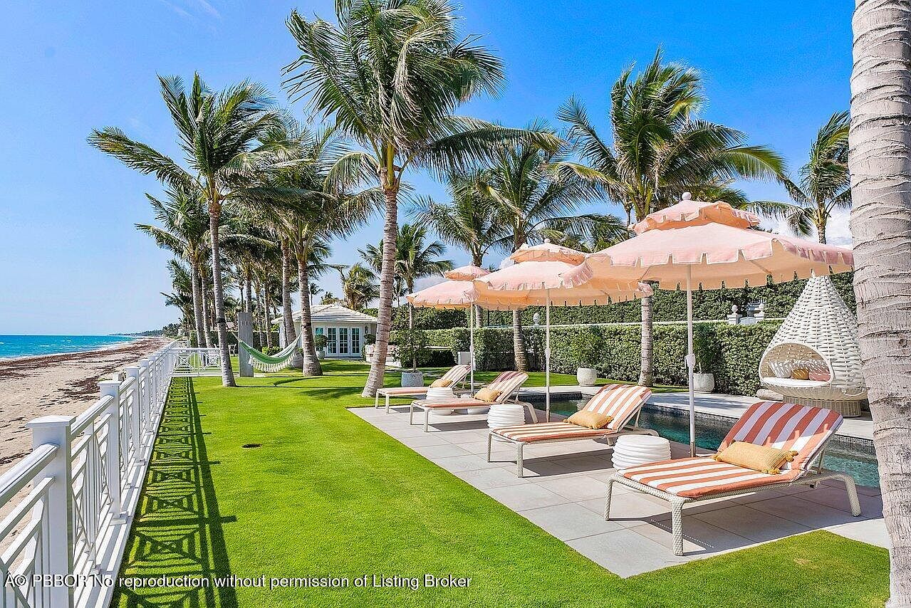 This exterior shot showcases a luxurious pool area with striped lounge chairs and peach-colored umbrellas, set against a backdrop of palm trees and a glimpse of the ocean. A white fence borders the lush green lawn, leading to a charming gazebo in the distance, creating an inviting and serene atmosphere perfect for relaxation and entertainment.