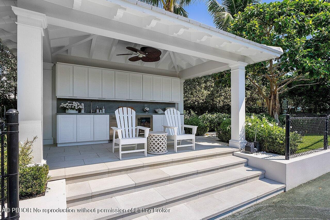 This image showcases a covered outdoor patio area, featuring white columns and a matching white roof structure. The patio includes built-in cabinetry with a countertop, two white Adirondack chairs, and a decorative side table. The space is accessed by stone steps, and lush greenery surrounds the area, creating a serene and inviting atmosphere.