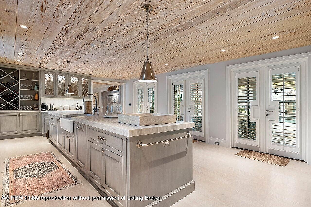 This is a well-lit kitchen featuring a large central island with a farmhouse sink and light-colored countertops. The kitchen includes custom cabinetry with glass-fronted upper cabinets and a built-in wine rack. French doors with shutters provide natural light and access to the outside, while pendant lights hang above the island, creating a warm and inviting atmosphere.