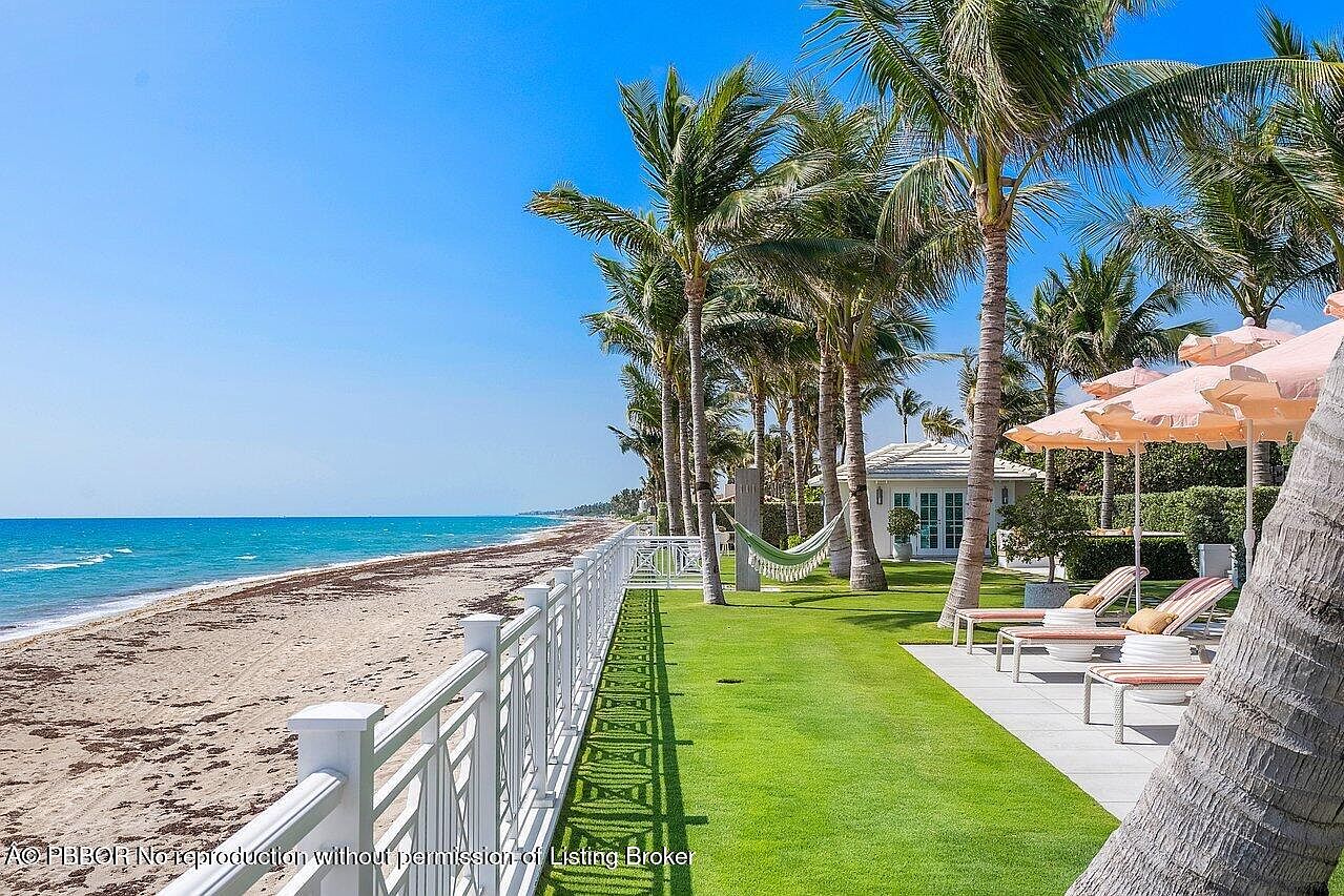 This image showcases a luxurious beachfront property with a meticulously maintained yard. Palm trees line the property, providing shade and a tropical ambiance. A white fence separates the yard from the sandy beach, and lounge chairs with umbrellas offer a relaxing space to enjoy the ocean view.